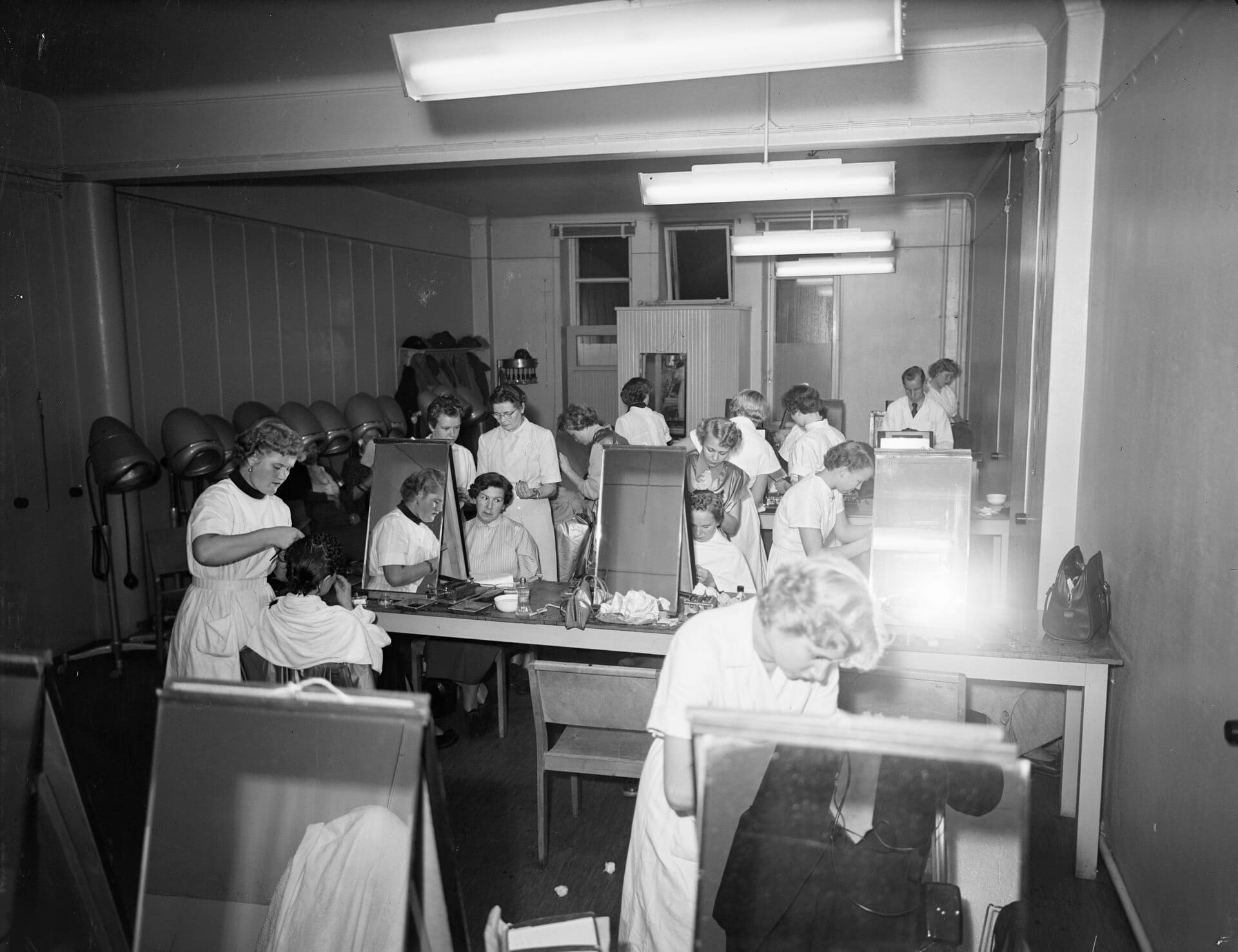 Students and clients in a hairdressing training room with workstations and hair dryers (black-and-white photograph, 1953)