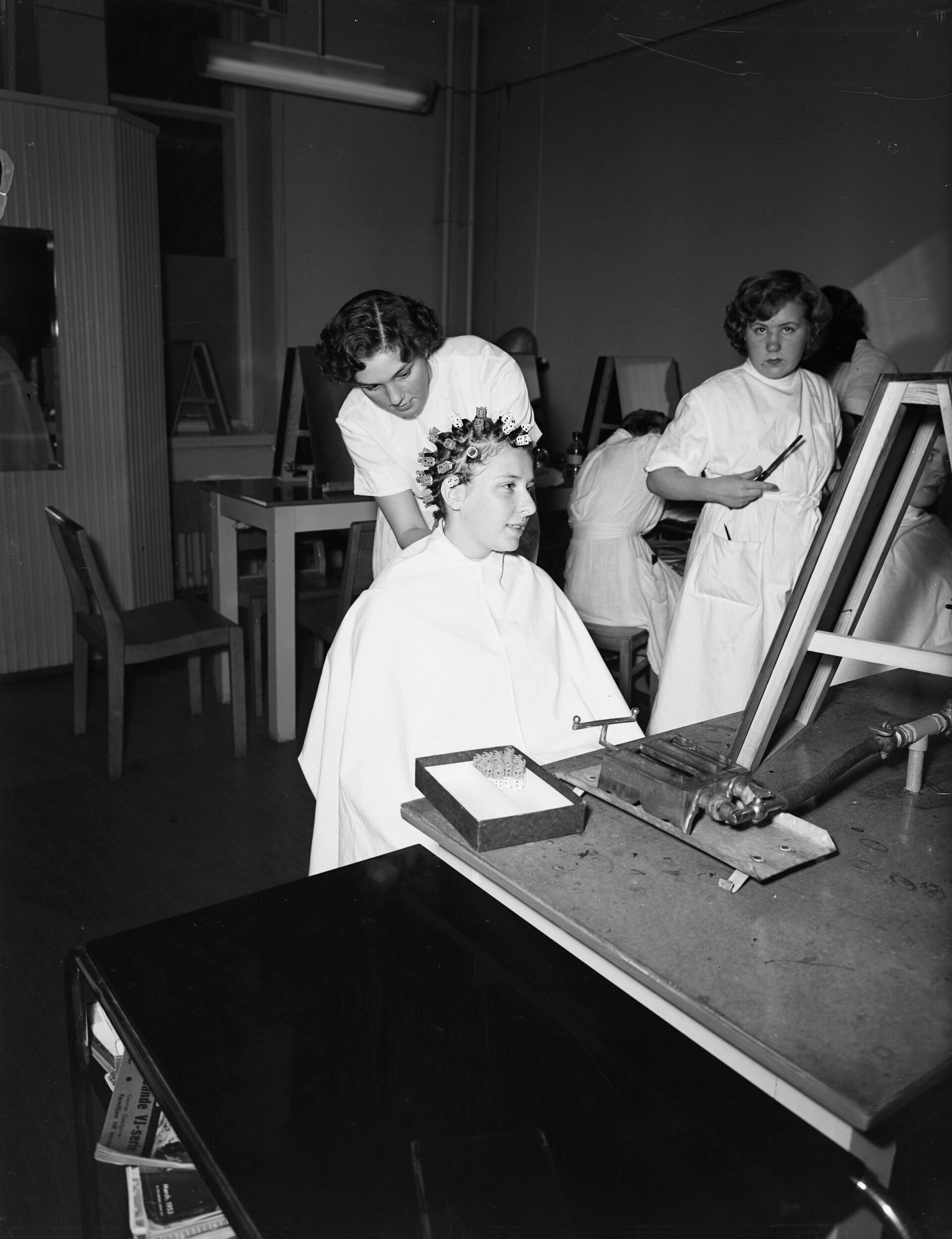 A hairdressing student setting a client's hair in rollers while another student looks on (black-and-white photograph, 1953)