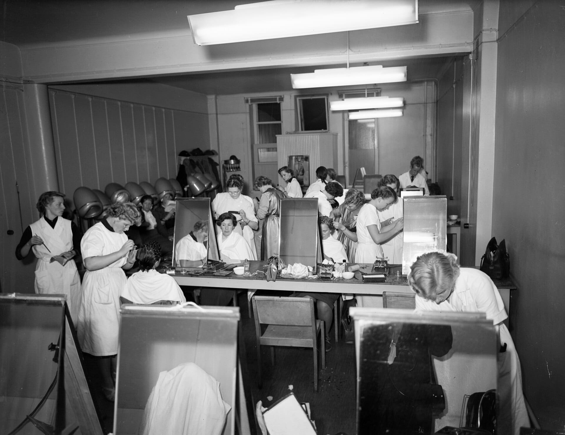 Students at work in a large vocational hairdressing and barbering training room, with rows of stations and mirrors (black-and-white photograph, 1953)